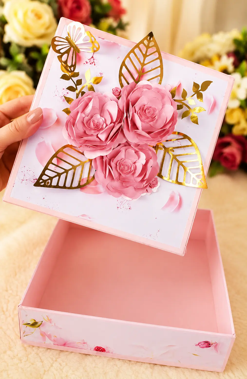 Caixa cartonada personalizada rosa com tampa decorada com flores de papel e folhas douradas, aberta mostrando o interior